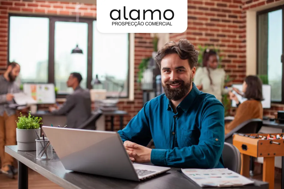 Homem sorrindo sentado à mesa com notebook em escritório moderno, com equipe trabalhando ao fundo e logotipo da Álamo Prospecção Comercial no topo da imagem.