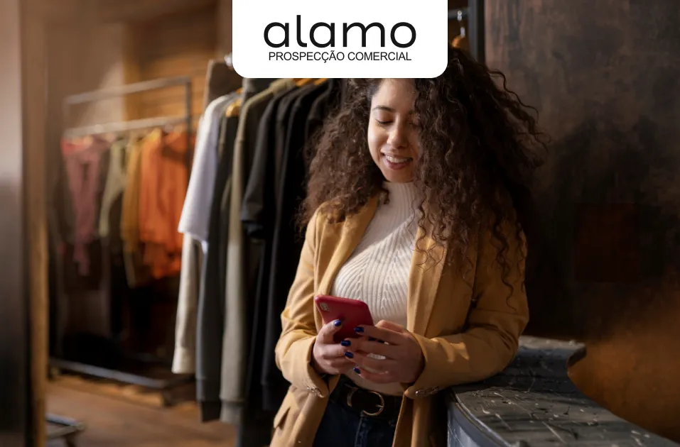 Fotografia de uma mulher jovem com cabelos cacheados, sorrindo enquanto utiliza um smartphone. Ela está em uma loja de roupas, encostada em um balcão, com araras de vestuário visíveis ao fundo. Na parte superior central da imagem, o logotipo da "Alamo Prospecção Comercial" está sobreposto em um retângulo de cantos arredondados.